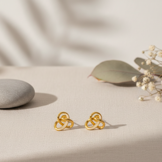 Gold knot earrings on a beige surface with a stone and dried flowers.