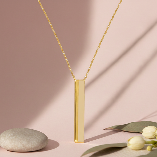 Gold necklace with a vertical bar pendant on a pink background with a stone and leaves.
