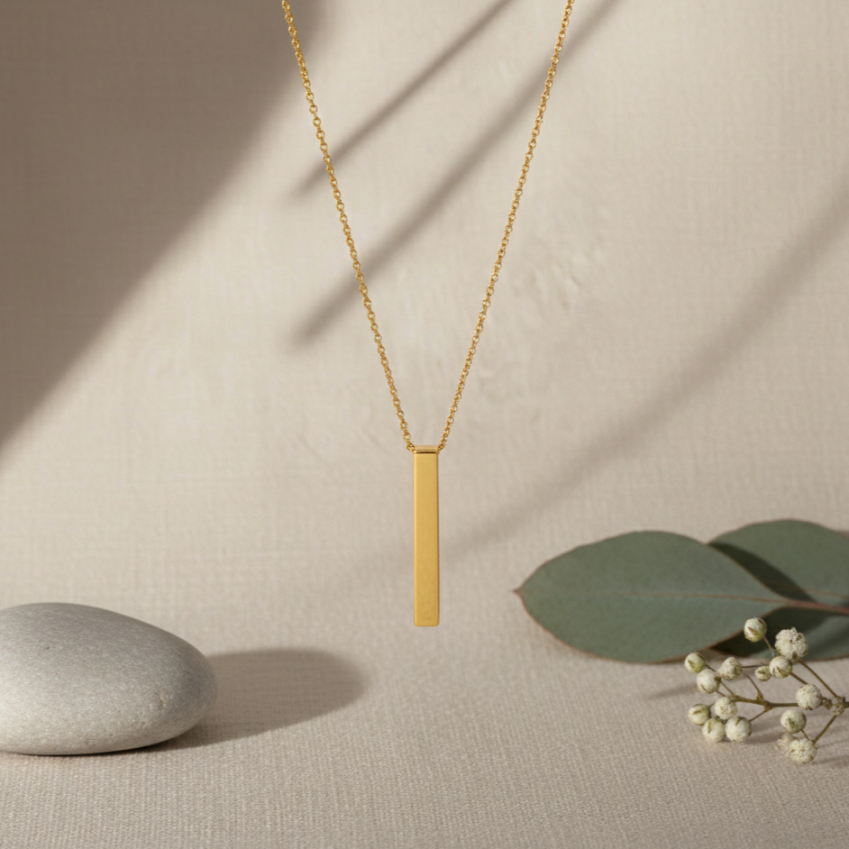 Gold necklace with a vertical bar pendant on a beige background with a stone and leaves.
